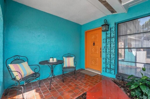 Una habitación con paredes turquesa y suelo de baldosas rojas, una pequeña mesa redonda, dos sillas de metal y una ventana con reja.