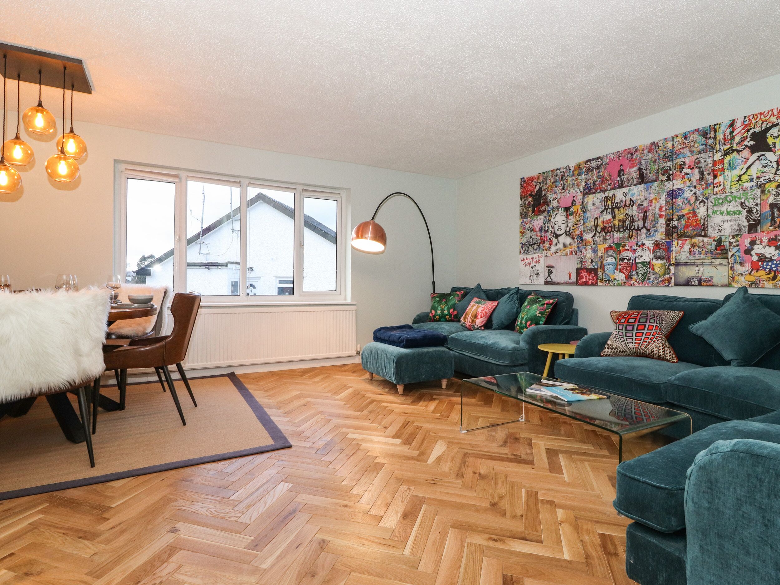 Ein modernes Wohnzimmer mit Fischgrätparkett, smaragdgrünen Sofas, einem Glastisch und einem großen, abstrakten Kunstwerk an der Wand.