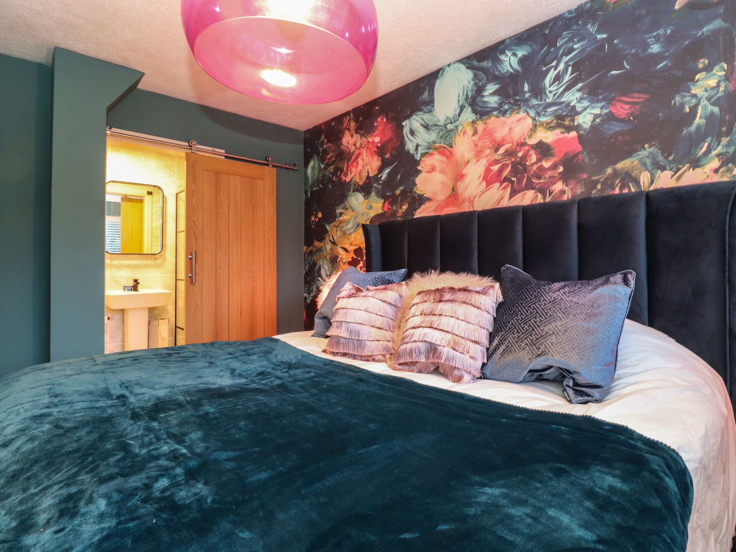 A bedroom with a bed, a headboard, a floral wall, a mirror, and a door.