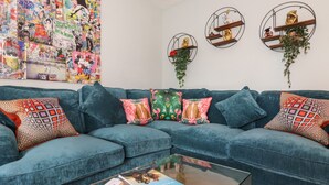 Un salon moderne avec un canapé d’angle bleu, des coussins colorés, une table basse en verre et une œuvre d’art murale.
