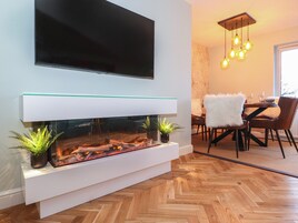 Uma sala de estar moderna com uma televisão de tela plana fixada na parede, uma lareira com frente de vidro e lenha, e uma área de jantar com um lustre.