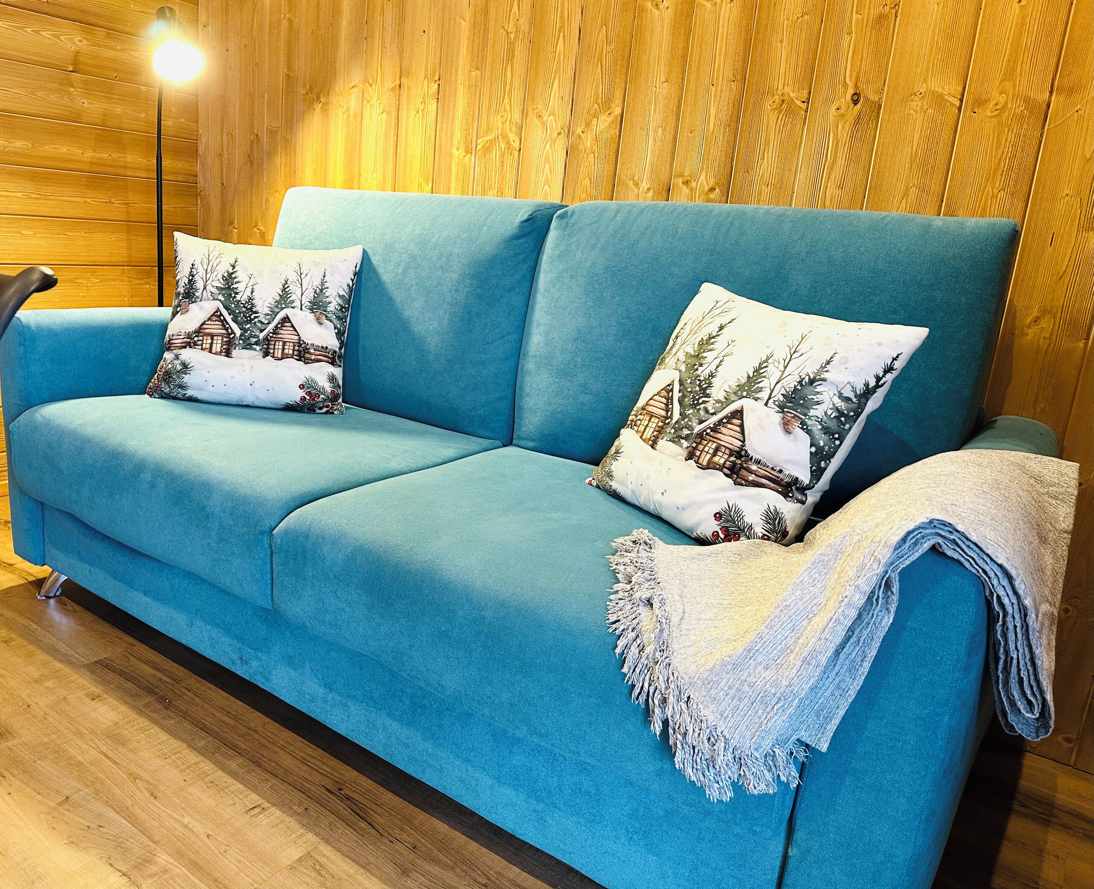 Un sofá color turquesa con dos almohadones decorativos y una manta, sobre piso de madera.