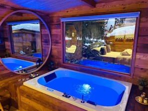 Interior de una cabaña de madera con una bañera grande, un espejo y una ventana con vistas nevadas.