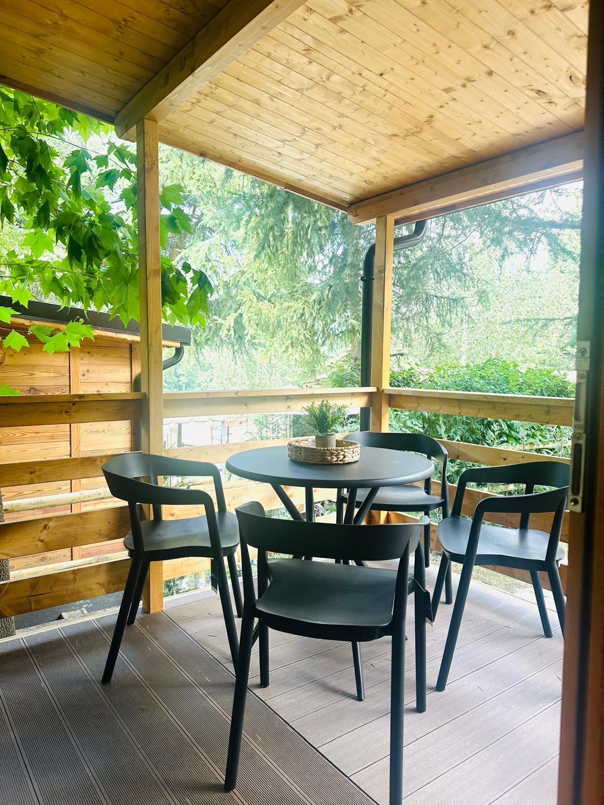 A covered patio with a wooden ceiling, a round table with chairs, and a view of greenery outside.