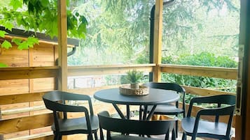 A covered patio with a wooden ceiling, a round table with chairs, and a view of greenery outside.
