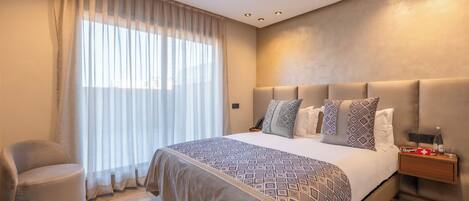 A modern bedroom with a large bed, a bedside table with a bottle, and a chair.