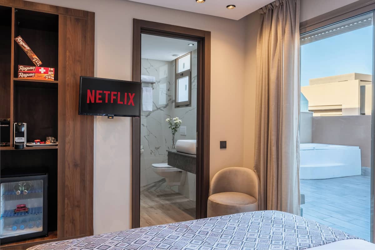 A modern hotel room with a large bed, a television displaying 'NETFLIX', a small seating area, and a view of a pool and building outside.