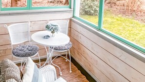 A cozy corner with a round table and wicker chairs, large windows with green frames, and string lights.