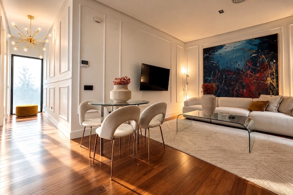 A modern living room with a glass coffee table, white chairs, a sofa, a flat-screen TV, and a large abstract painting.