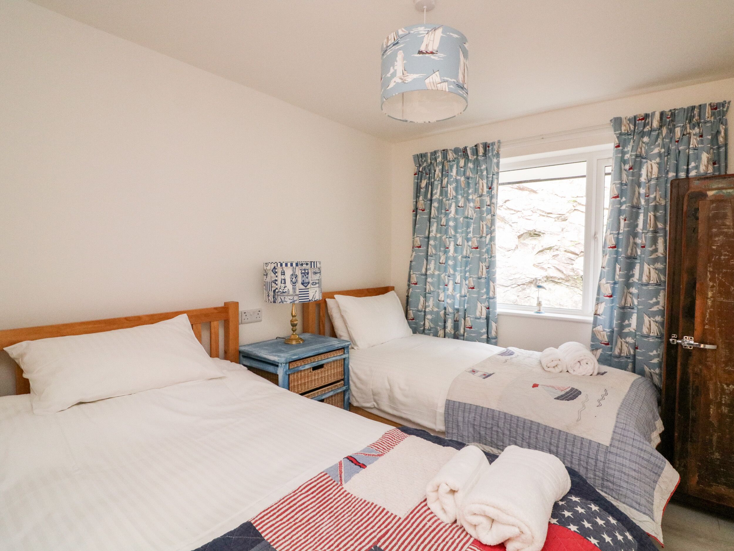 A bedroom with two beds, a nightstand, a lamp, and curtains with a sailboat pattern.