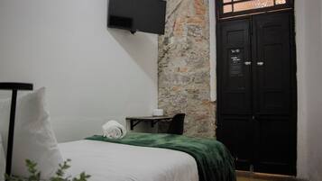 A hotel room with a bed, a flat-screen TV mounted on the wall, a stone wall, a black door, and a small table with a chair.