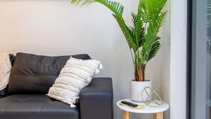 A modern living room with a black leather sofa, a white throw blanket, a potted plant, a small round table with a remote control, and a large window. - Botanic Bliss Bungalow: 1 bed loft (Christchurch)