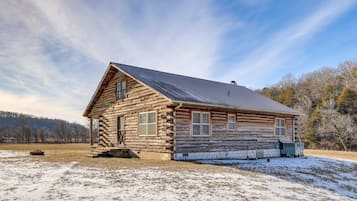 A log cabin with a metal roof, situated in a snowy field with trees in the background.