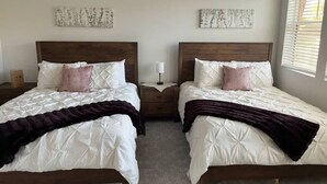Une chambre avec deux lits, chacun doté d’une tête de lit en bois, de draps blancs et d’un plaid foncé. On y trouve deux tables de chevet avec des lampes, ainsi que des œuvres d’art accrochées aux murs.