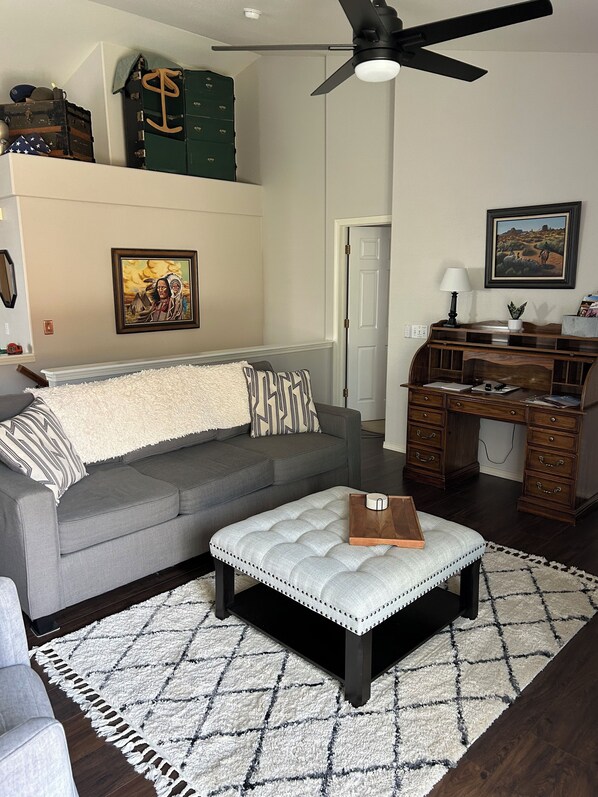 A living room with a gray sofa, a tufted ottoman, a wooden desk, and a ceiling fan. - 4 Bedroom/3 Bath - Spacious Briargate Ranch Home with Office and WiFi near USAFA (Colorado Springs)