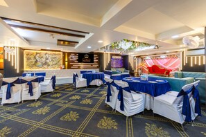 A banquet hall with round tables, chairs with blue sashes, a stage with floral decorations, and a ceiling with recessed lighting.