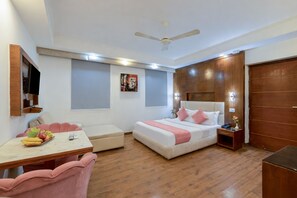 A hotel room with a bed, a desk with fruit, a chair, a TV, and a ceiling fan. - The Royal Galaxy (New Delhi)