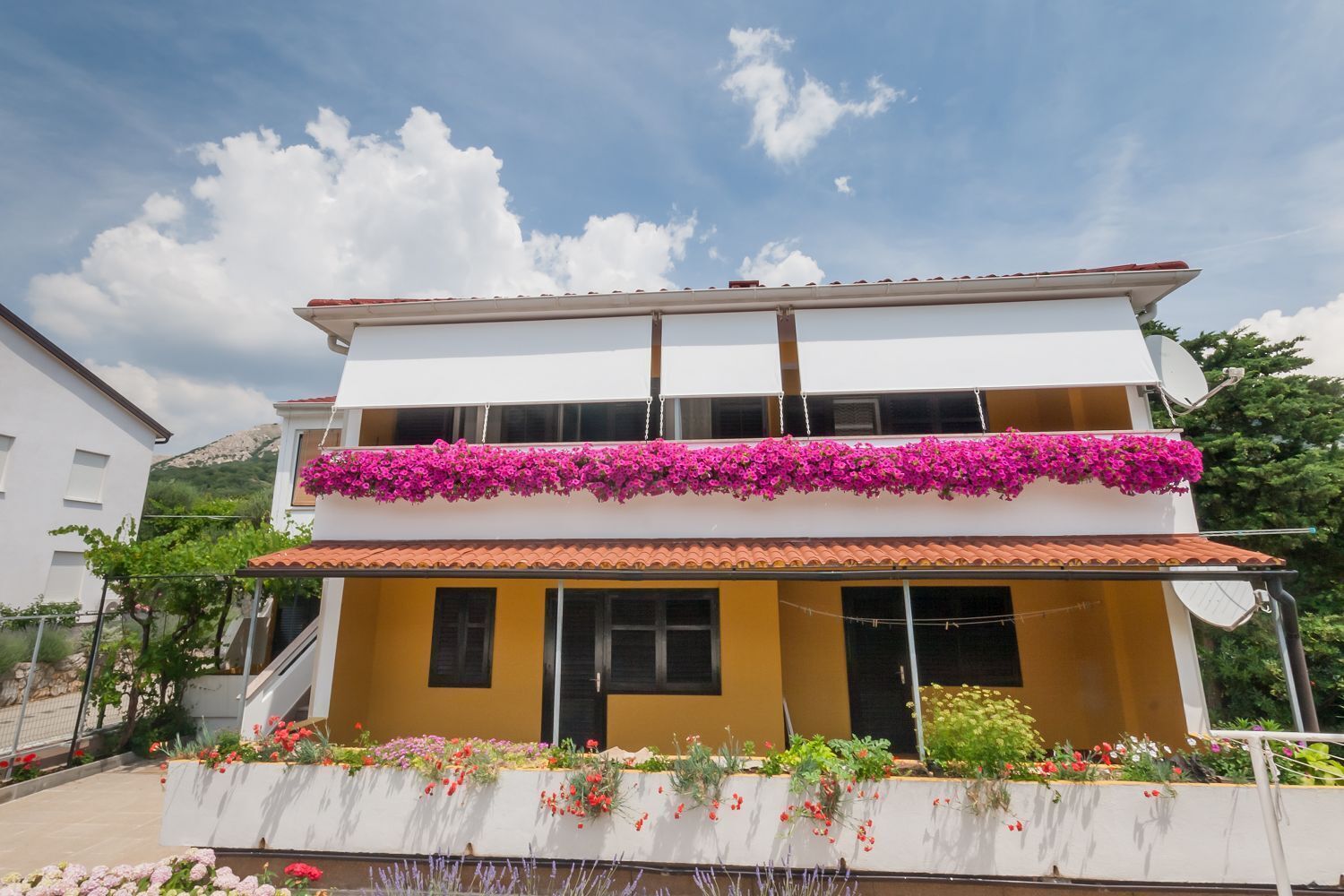 A two-story building with a balcony adorned with pink flowers, a tiled roof, and a white facade.