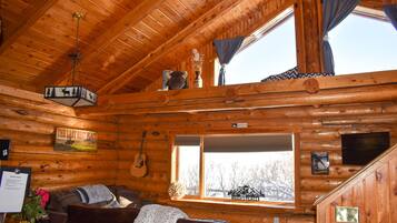 A log cabin interior with a wooden ceiling, a large window, a couch, a guitar, and a sign reading "The Last Best Place".