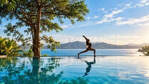 Una persona realiza una postura de yoga en la cubierta de una piscina, con vistas a un cuerpo de agua y montañas al fondo.