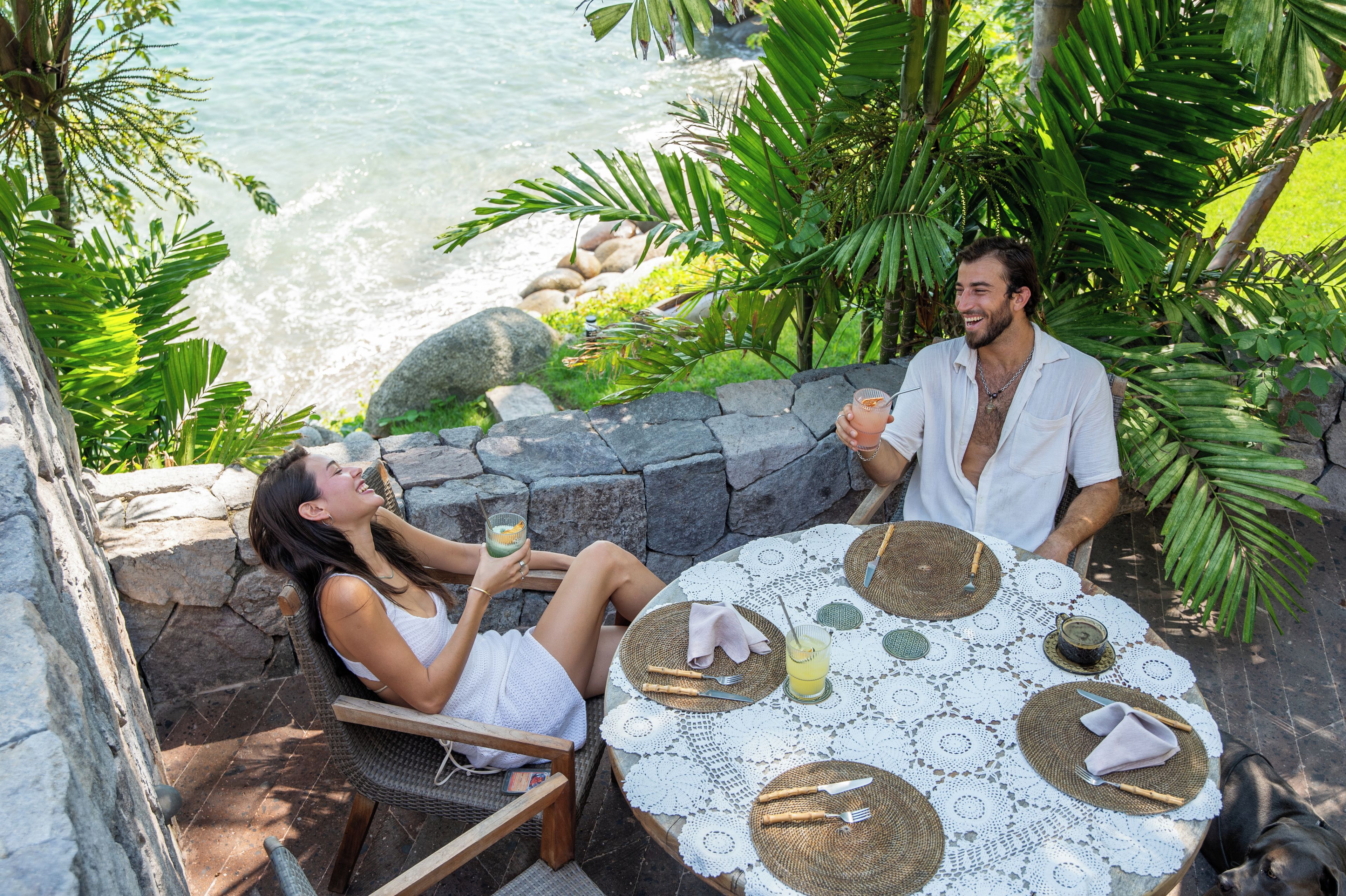 A couple enjoying a drink at an outdoor table with a view of the ocean, surrounded by tropical plants.