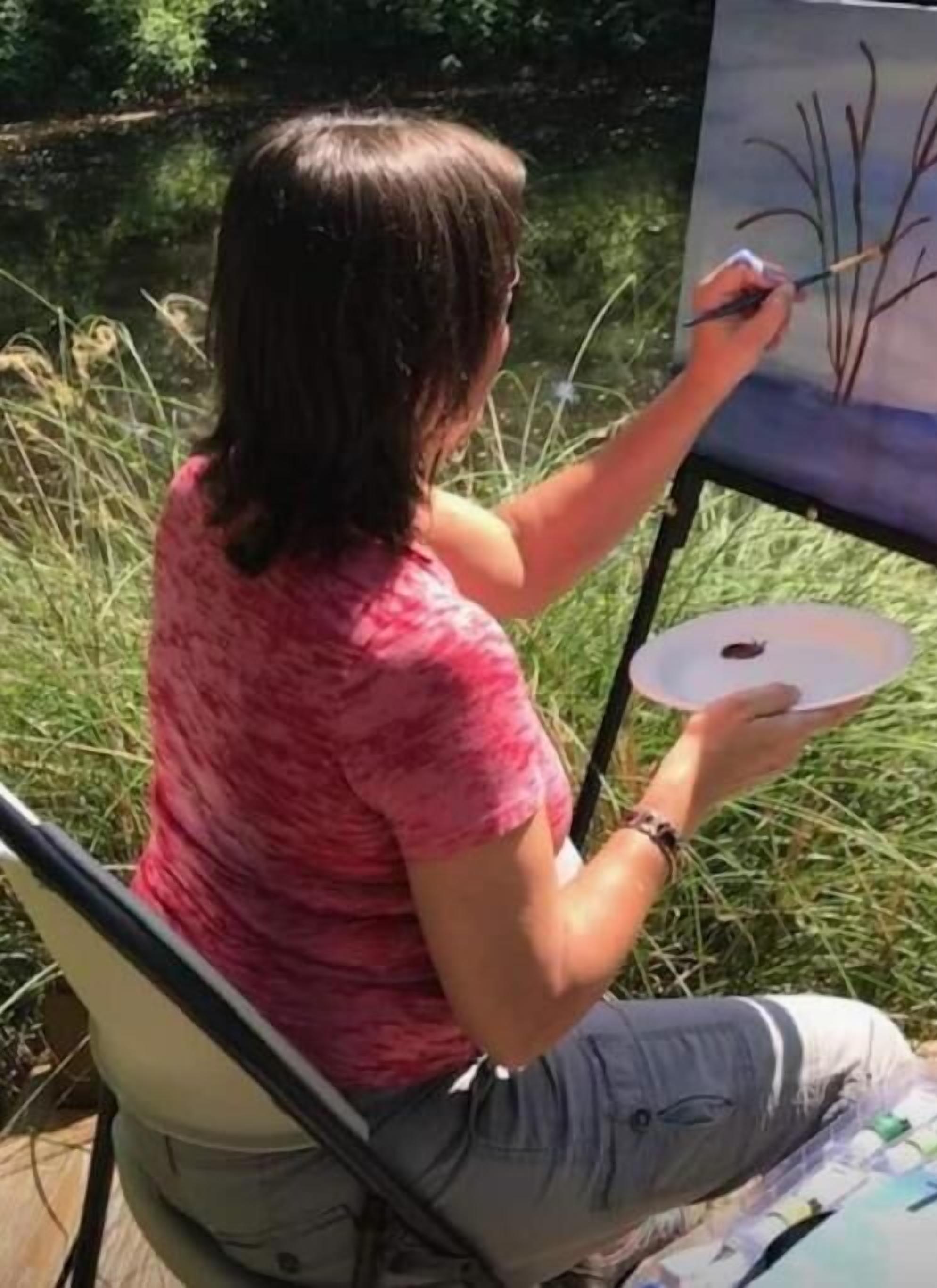 Una persona pintando al aire libre, sentada a una mesa con un lienzo, en un entorno natural.