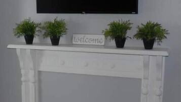 A white fireplace with a 'welcome' sign, flanked by three potted plants, and a flat-screen TV mounted above it.