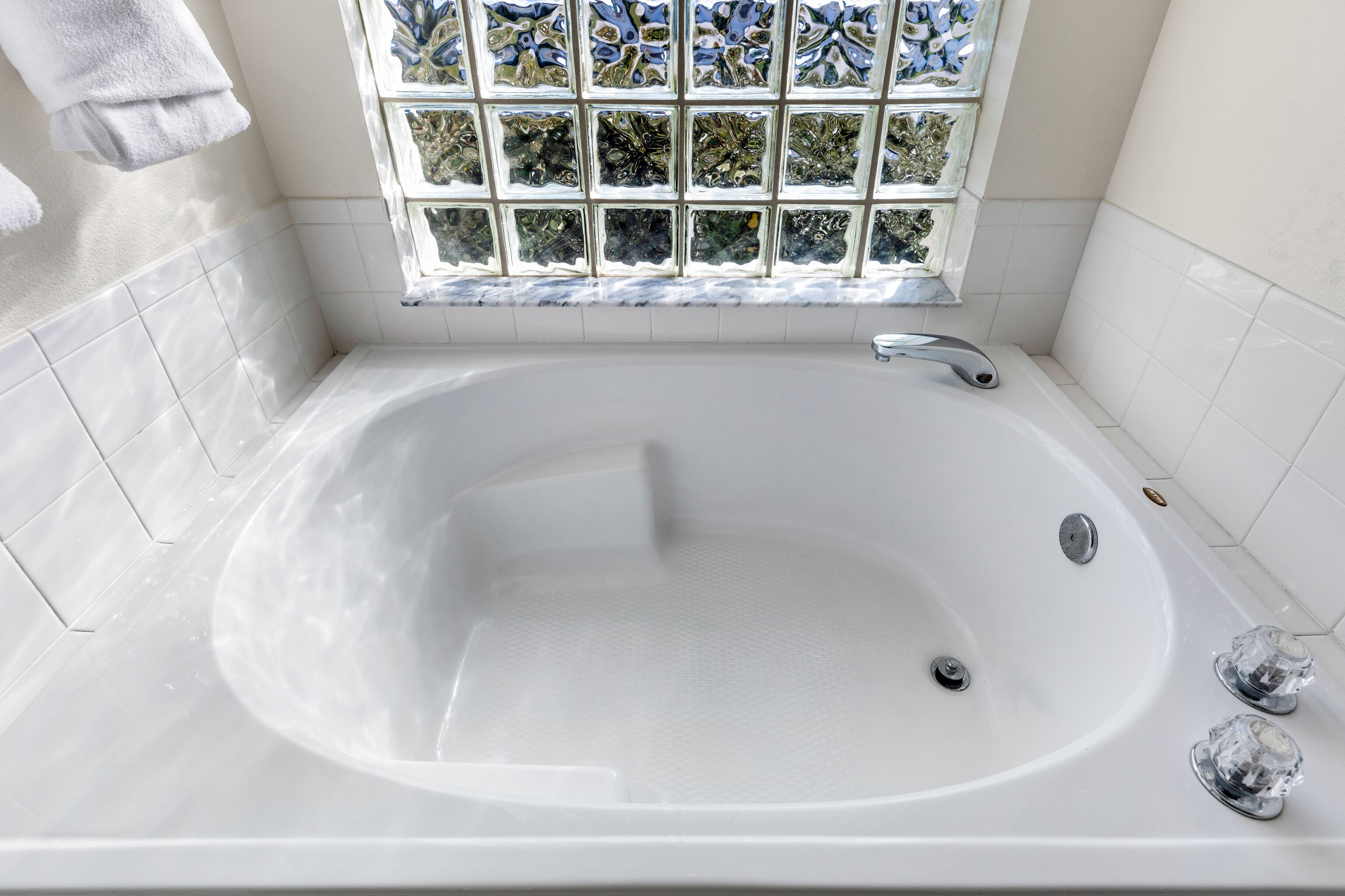 A large, white bathtub with a glass window above it, white tiled walls, and a towel hanging on the wall.