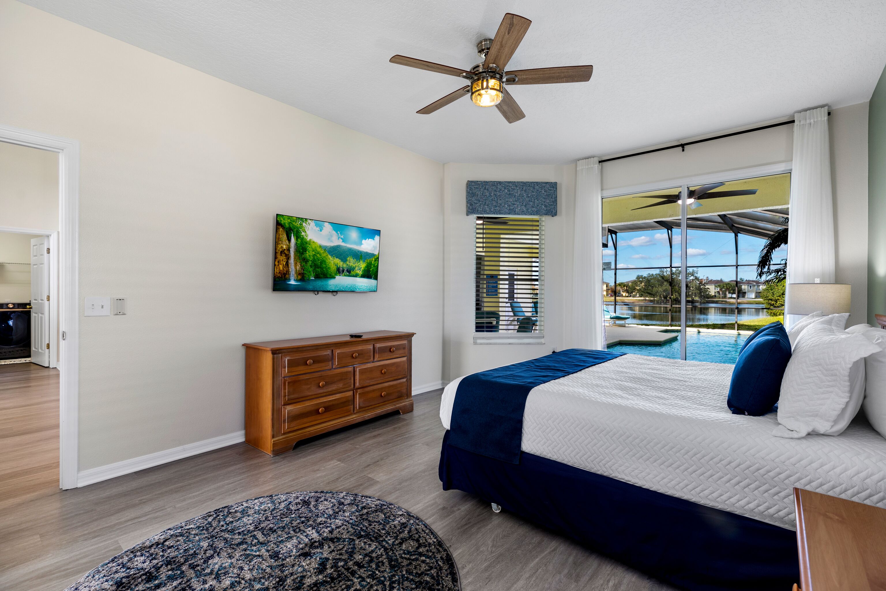 A bedroom with a bed, a ceiling fan, a TV, a window with blinds, and a view of a swimming pool.