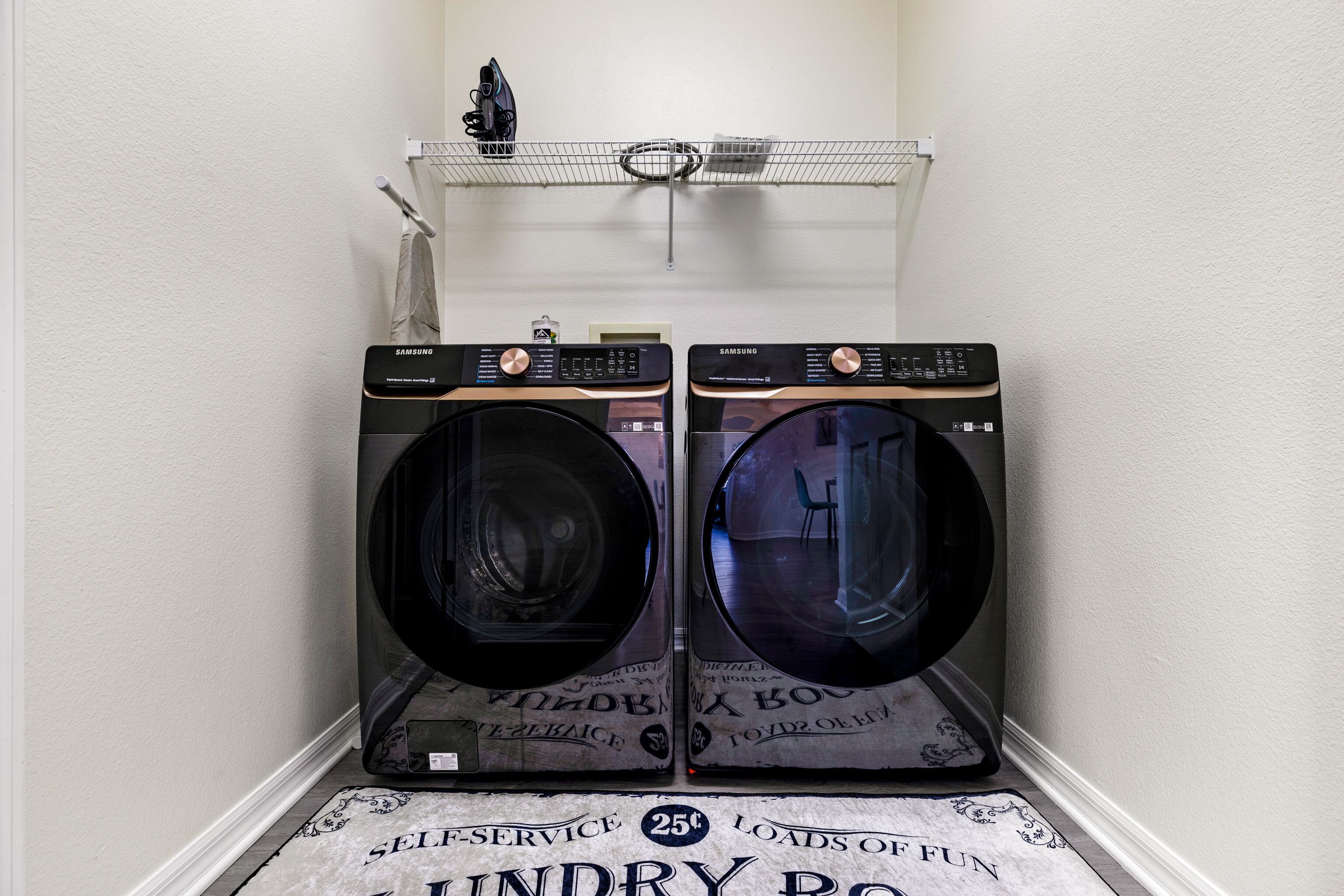 A laundry room with two Samsung washing machines, a shelf above with an iron and a basket, and a self-service sign on the floor.