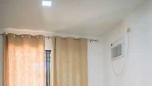 Een enkel bed met een houten hoofdbord, beige gordijnen en een witte muur met een raam en een airconditioning.