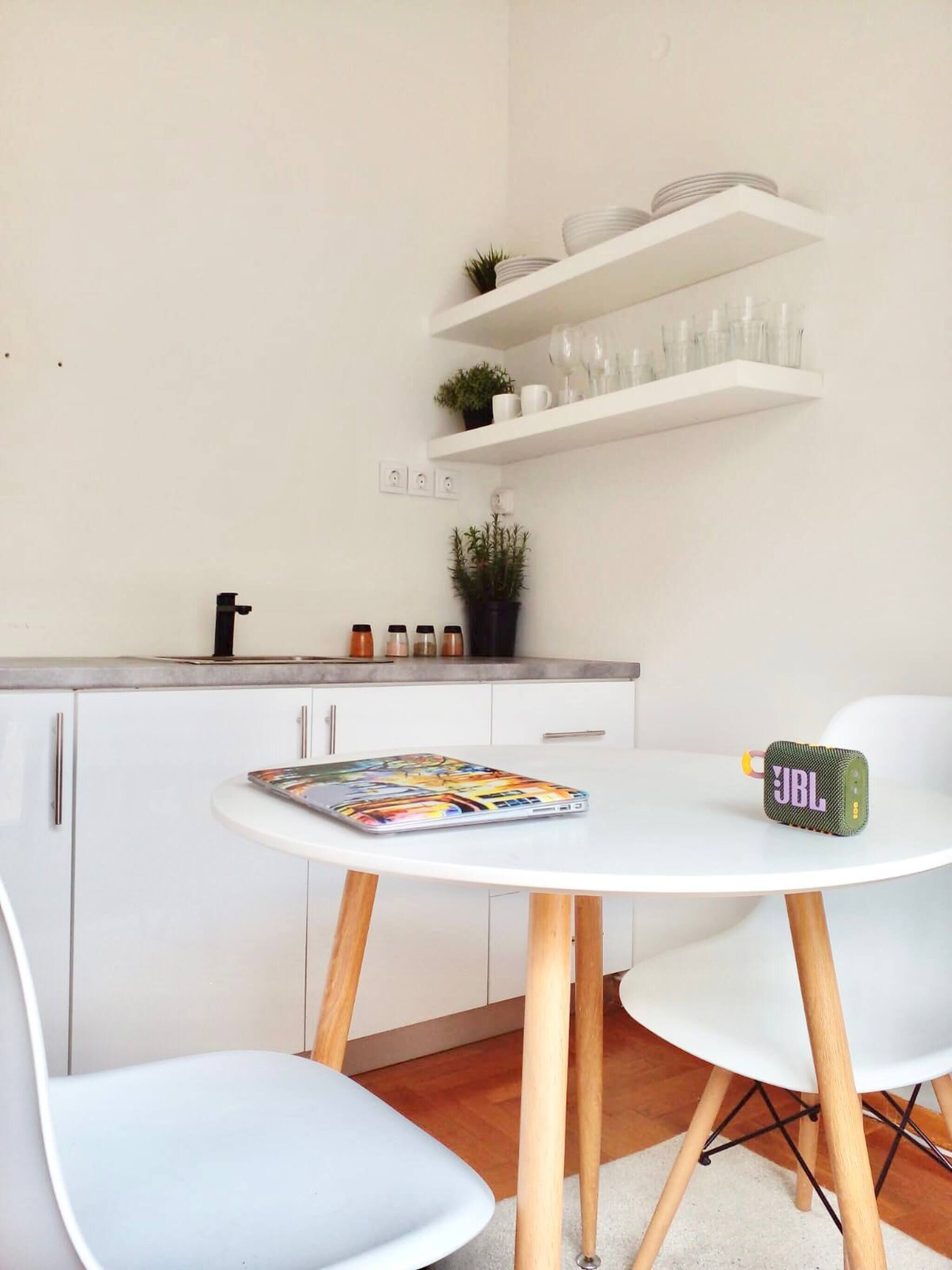 Une cuisine moderne avec des armoires blanches, un plan de travail blanc et une table ronde entourée de chaises. Des étagères au-dessus du plan de travail servent à ranger la vaisselle et les verres, et une petite plante est posée sur l’une de ces étagères.