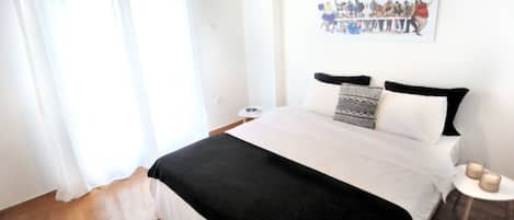 Quarto com cama, cortinas brancas, um quadro na parede e uma pequena mesa de cabeceira com velas.