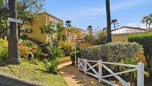 Un edificio amarillo con cerca blanca, rodeado de abundante vegetación y palmeras.