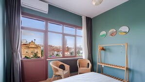 Habitación de hotel con un ventanal grande, una cama, dos sillas de mimbre y un mueble estante de madera.