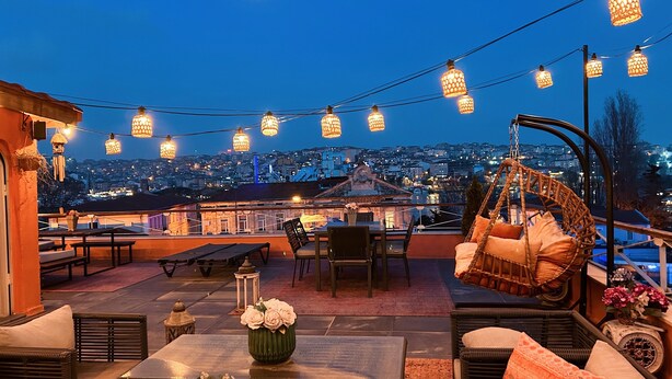 Terraza al aire libre con muebles de mimbre, luces colgantes y vistas a la ciudad al anochecer.