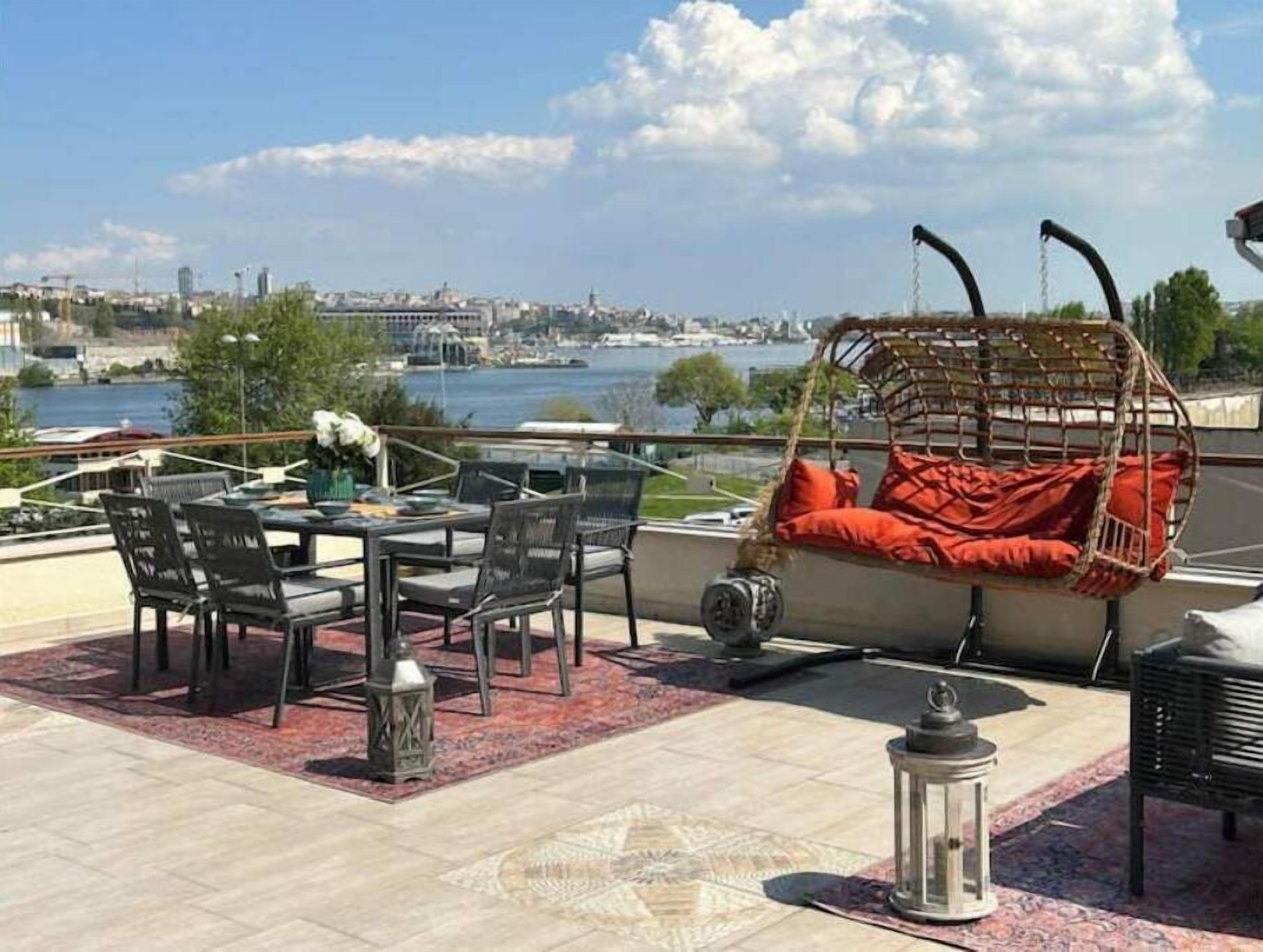 A rooftop terrace with a dining table, chairs, a hanging chair with red cushions, and a view of a cityscape and water.