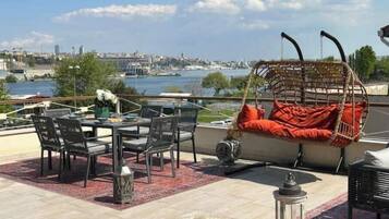 A rooftop terrace with a dining table, chairs, a hanging chair with red cushions, and a view of a cityscape and water.