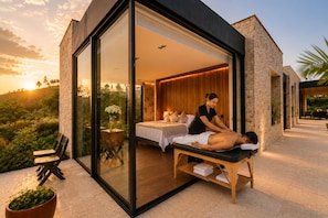 Une salle de spa moderne équipée d’une table de massage, d’un lit et d’un mur en verre donnant sur l’extérieur.