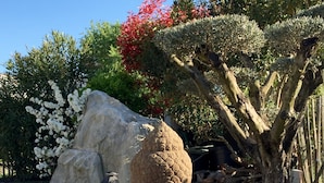 A garden with a large rock, a tree with red flowers, and a stone sculpture.