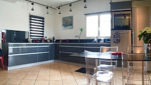 A modern kitchen with a central island, black and white cabinetry, a stainless steel refrigerator, and a dining area with clear glass chairs.