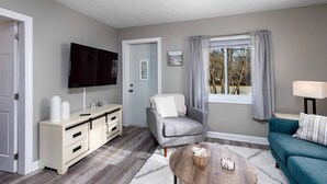 A living room with a TV, a grey armchair, a teal sofa, a wooden coffee table, and a white cabinet.