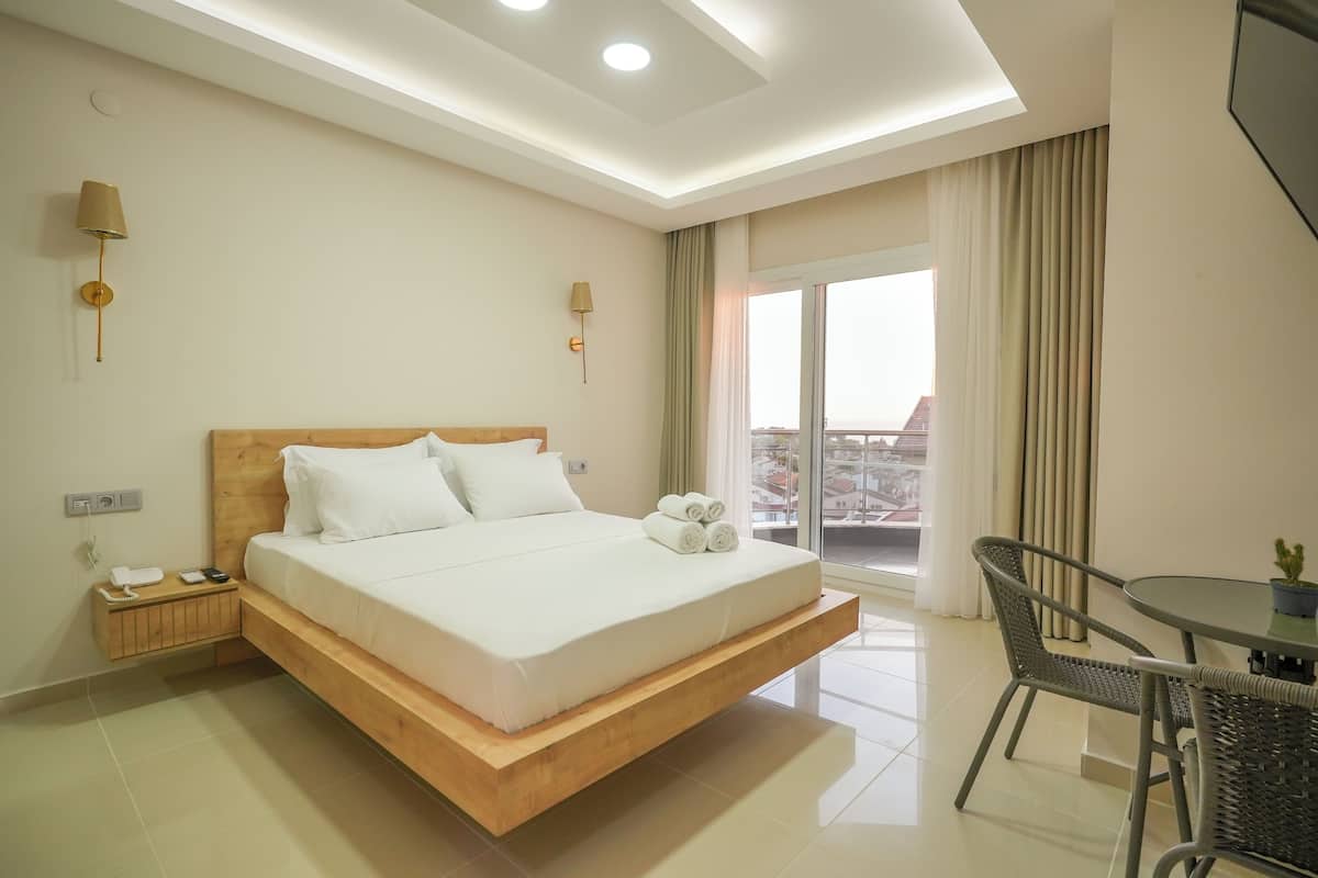 A modern hotel room with a wooden bed, a small bedside table with a phone and charger, a round dining table with chairs, and a balcony with curtains.