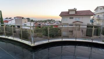 A pool area with a glass balustrade, surrounded by buildings and a sunset view.