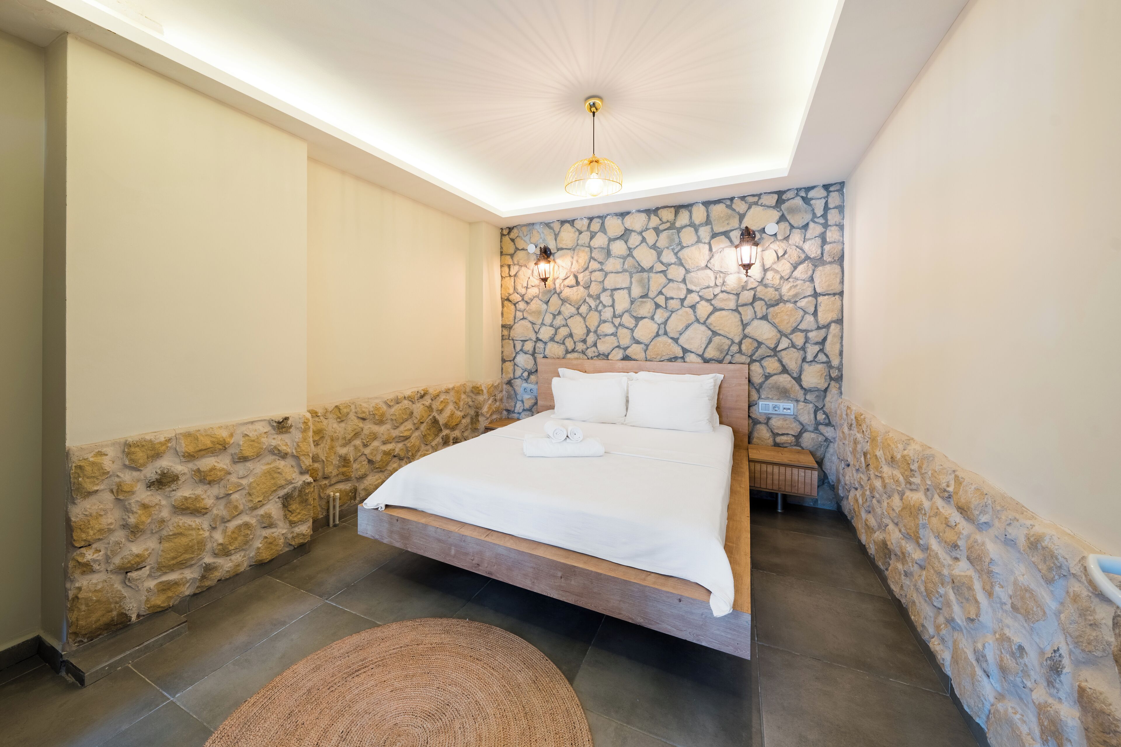 A bedroom with a stone wall, a bed with white linens, and two wall-mounted lights.