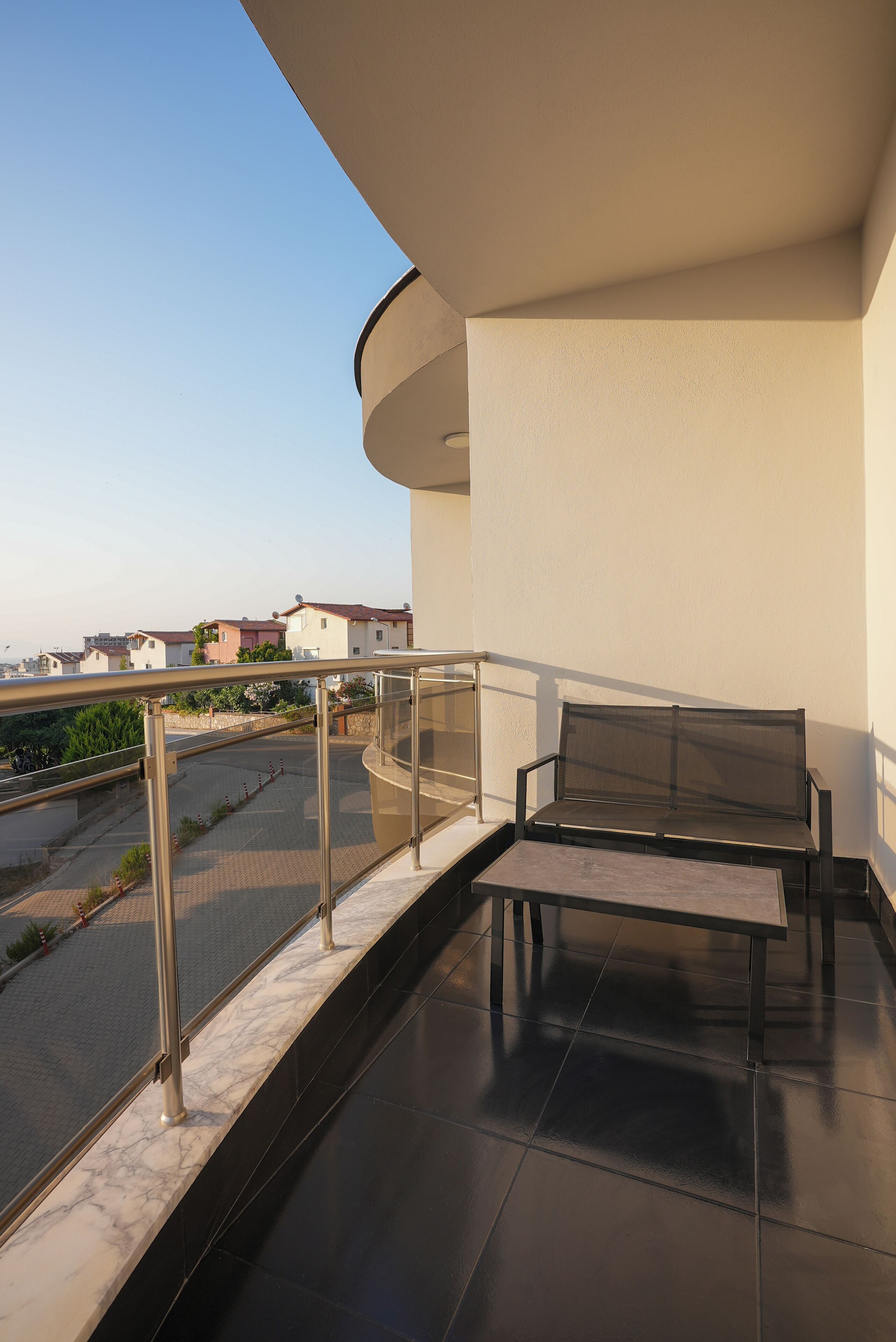 Un balcon doté d’un banc rembourré et d’une petite table, donnant sur un quartier résidentiel avec des maisons et une route.
