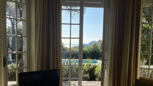 Un salon avec un téléviseur posé sur un meuble, un canapé et une vue sur une piscine et un espace verdoyant, visible à travers de grandes portes vitrées.