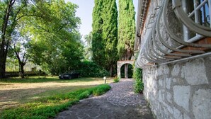 Un chemin de pierre menant à une entrée en arc de cercle, bordé d’une végétation luxuriante et de grands arbres.