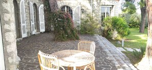 Un patio de piedra con una mesa y sillas de mimbre, rodeado de muros de piedra y ventanas de arco con contraventanas.