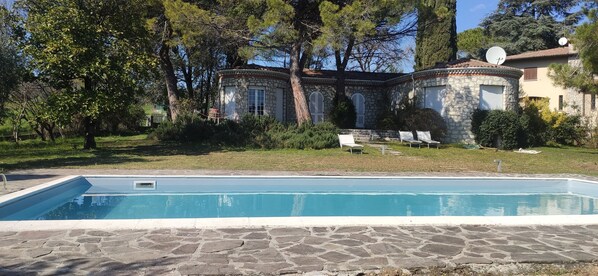 Una piscina de piedra con un suelo de piedra, rodeada de césped cuidado y árboles.